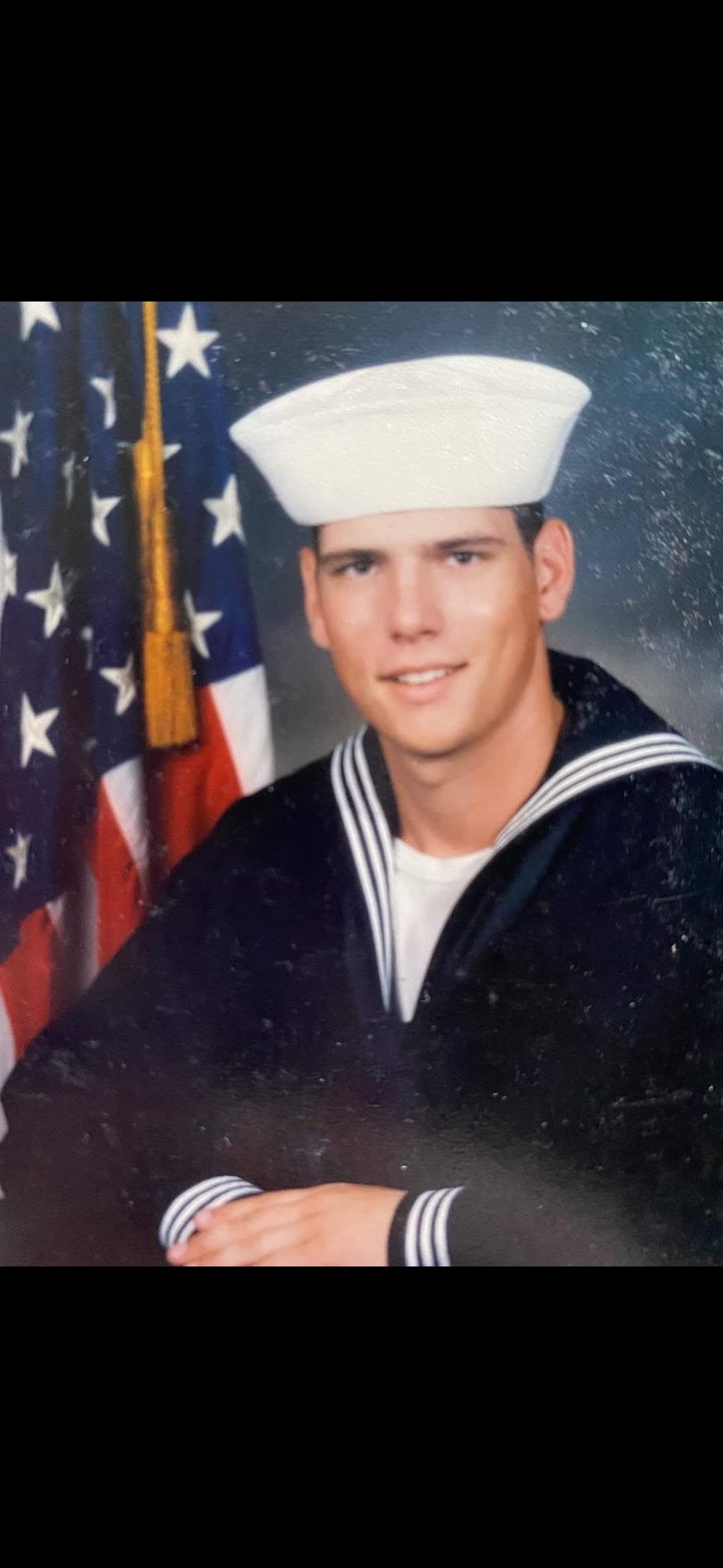 A vintage portrait of a young man in a US Navy uniform with a white sailor hat, seated in front of an American flag.